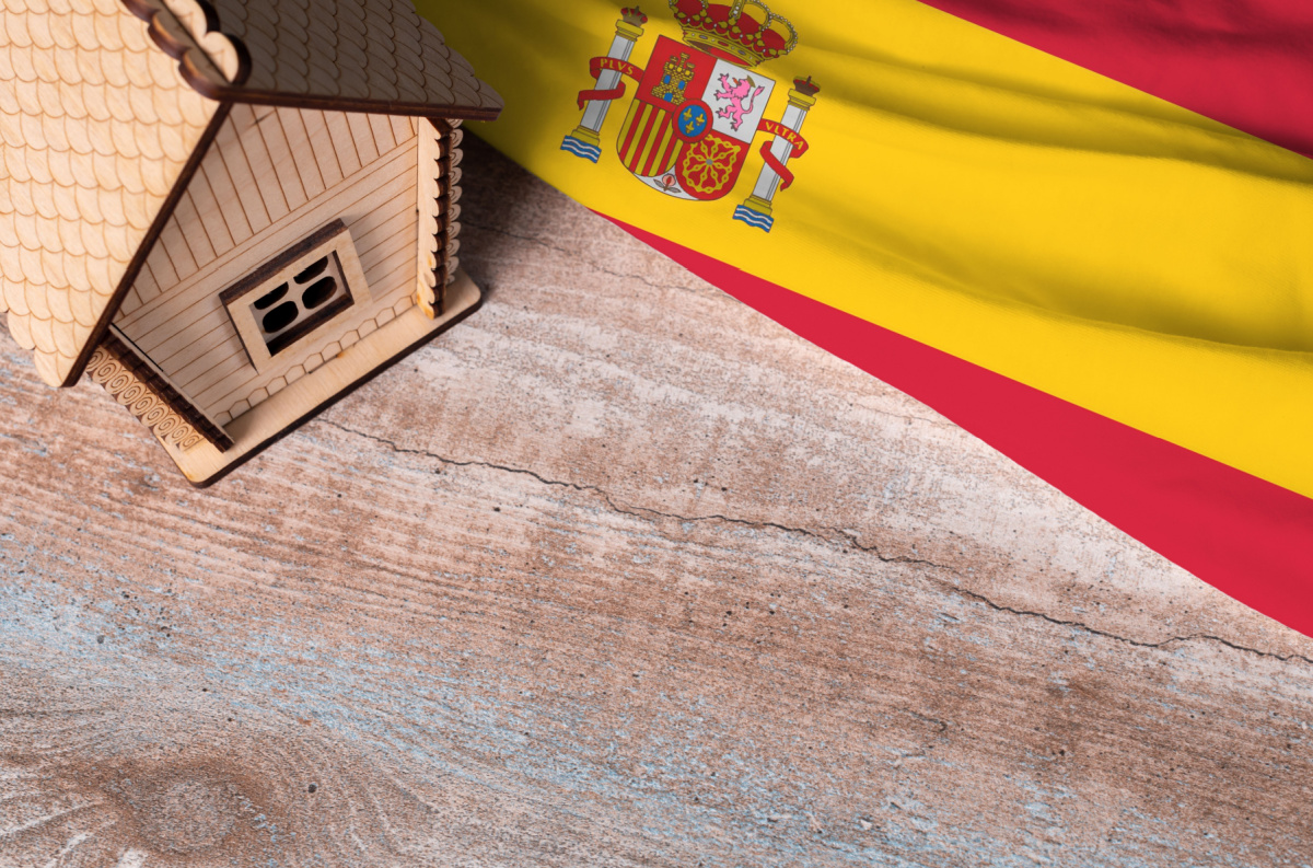 A wooden house model placed next to the Spanish flag on a rustic surface, symbolizing the process and requirements for relocating to Spain.